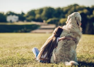 A child in a white shirt hugs a fluffy golden dog while sitting on the grass in an open field with trees in the background.