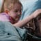 A large, relaxed brown dog laying in a hospital bed with a young girl. She is wearing a nasal cannula.
