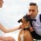 A bride and groom pet and comfort a dog dressed in a black bow tie. The dog happily licks the groom's cheek.