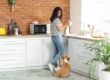 A young woman wearing a striped T-shirt and jeans drying a cup with a towel while looking down at a corgi next to her.