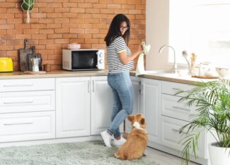 A young woman wearing a striped T-shirt and jeans drying a cup with a towel while looking down at a corgi next to her.