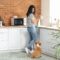 A young woman wearing a striped T-shirt and jeans drying a cup with a towel while looking down at a corgi next to her.