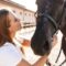 A smiling young woman stands outside and holds onto her horse's bridle as she looks up at the horse.
