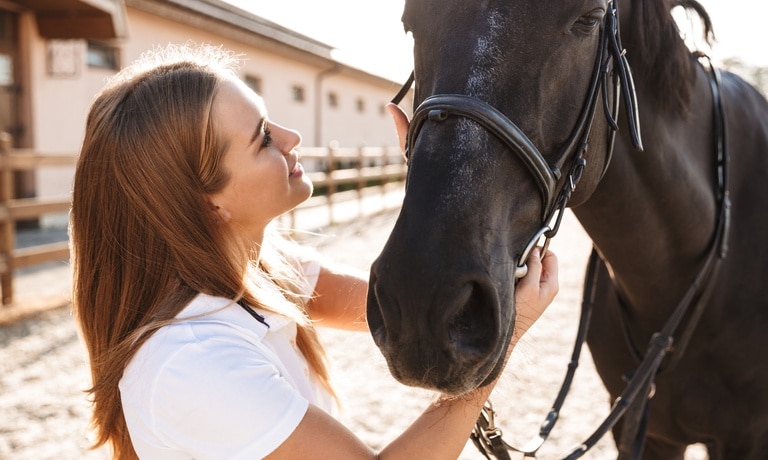 A smiling young woman stands outside and holds onto her horse's bridle as she looks up at the horse.