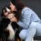 Woman in denim outfit kneeling and kissing a large black and brown dog with tongue out, sitting by a dark wall.