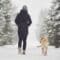 A person in a winter coat walking on a snowy trail with their dog next to them. Around them, trees are covered in snow.