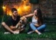 A couple plays with their dog in their backyard at night. String lights hang along the brick wall behind them.