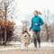 A woman wearing a light blue coat and gray leggings is jogging beside a large, fluffy dog. Snow lightly covers the ground.