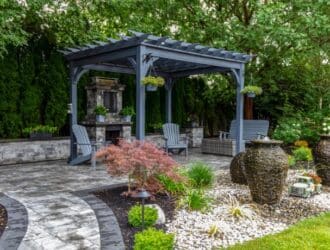 An outdoor space with a large grey pergola boasts ample seating, a stone fireplace, curved pavers, and decorative landscaping.
