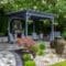 An outdoor space with a large grey pergola boasts ample seating, a stone fireplace, curved pavers, and decorative landscaping.