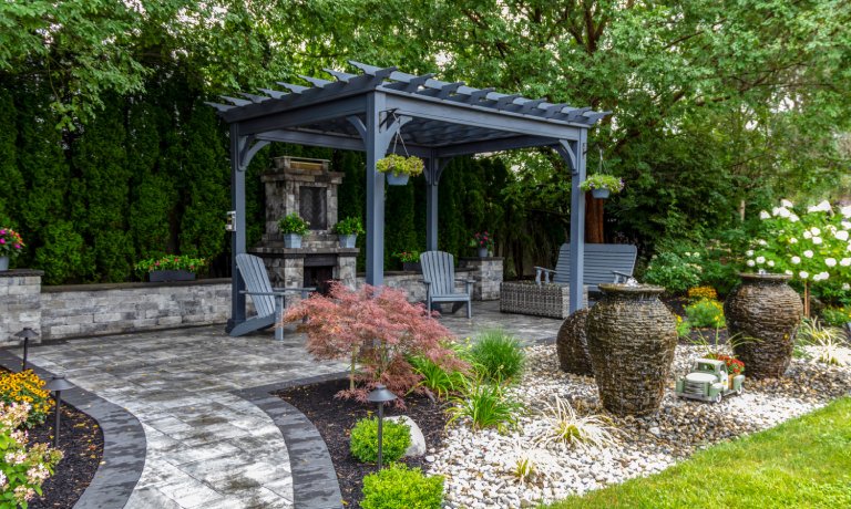 An outdoor space with a large grey pergola boasts ample seating, a stone fireplace, curved pavers, and decorative landscaping.