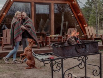 A couple wrapped in a blanket stands outside a tiny home with a brown dog. They have a fire going on a camp stove.