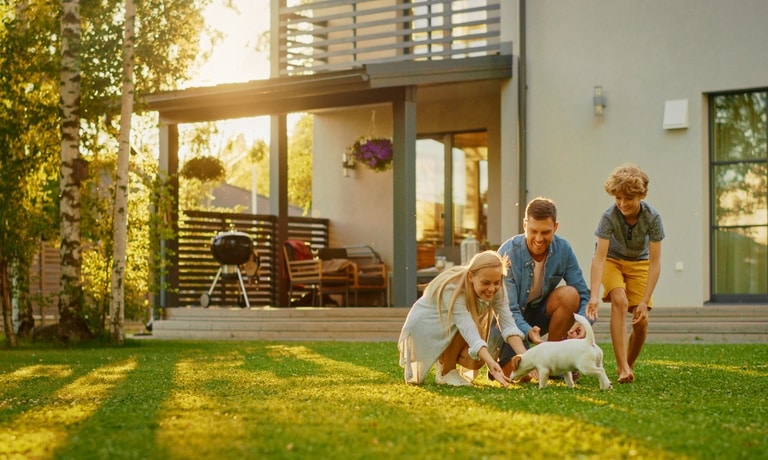 A smiling father, mother, and son play with a dog in the backyard of their home, as the sun shines in the background.