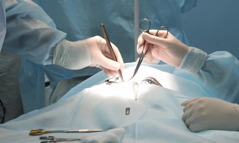 Close-up of a surgical procedure with two gloved professionals using instruments on a patient under bright lighting.