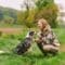 In a green field, a woman kneels beside a harnessed dog, hand raised to its nose near quiet autumn trees.