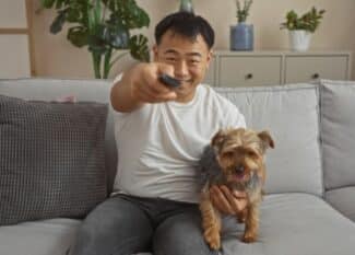 A man smiles as he holds his pet dog in one hand and a remote in the other. He leans over to turn on a television.