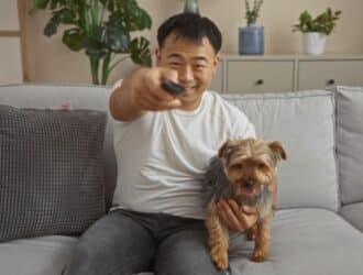 A man smiles as he holds his pet dog in one hand and a remote in the other. He leans over to turn on a television.