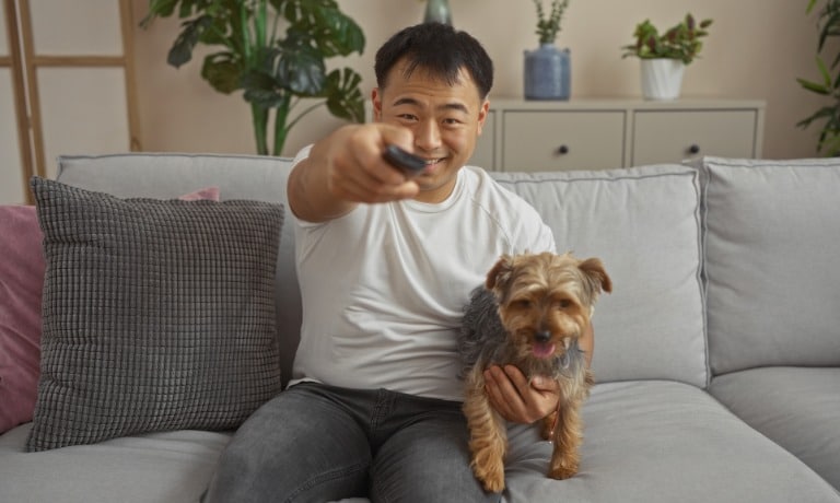 A man smiles as he holds his pet dog in one hand and a remote in the other. He leans over to turn on a television.