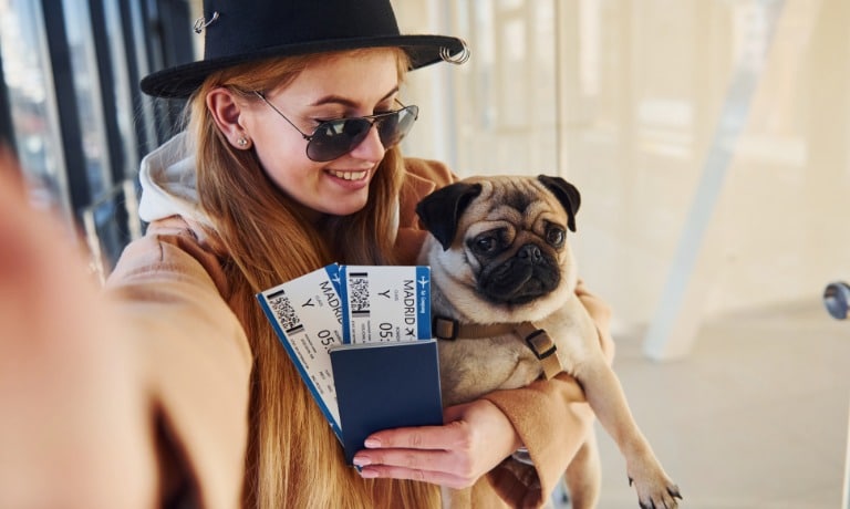 A woman takes a selfie while holding her pug in her arms. She also holds two airplane tickets to Madrid in her hand.